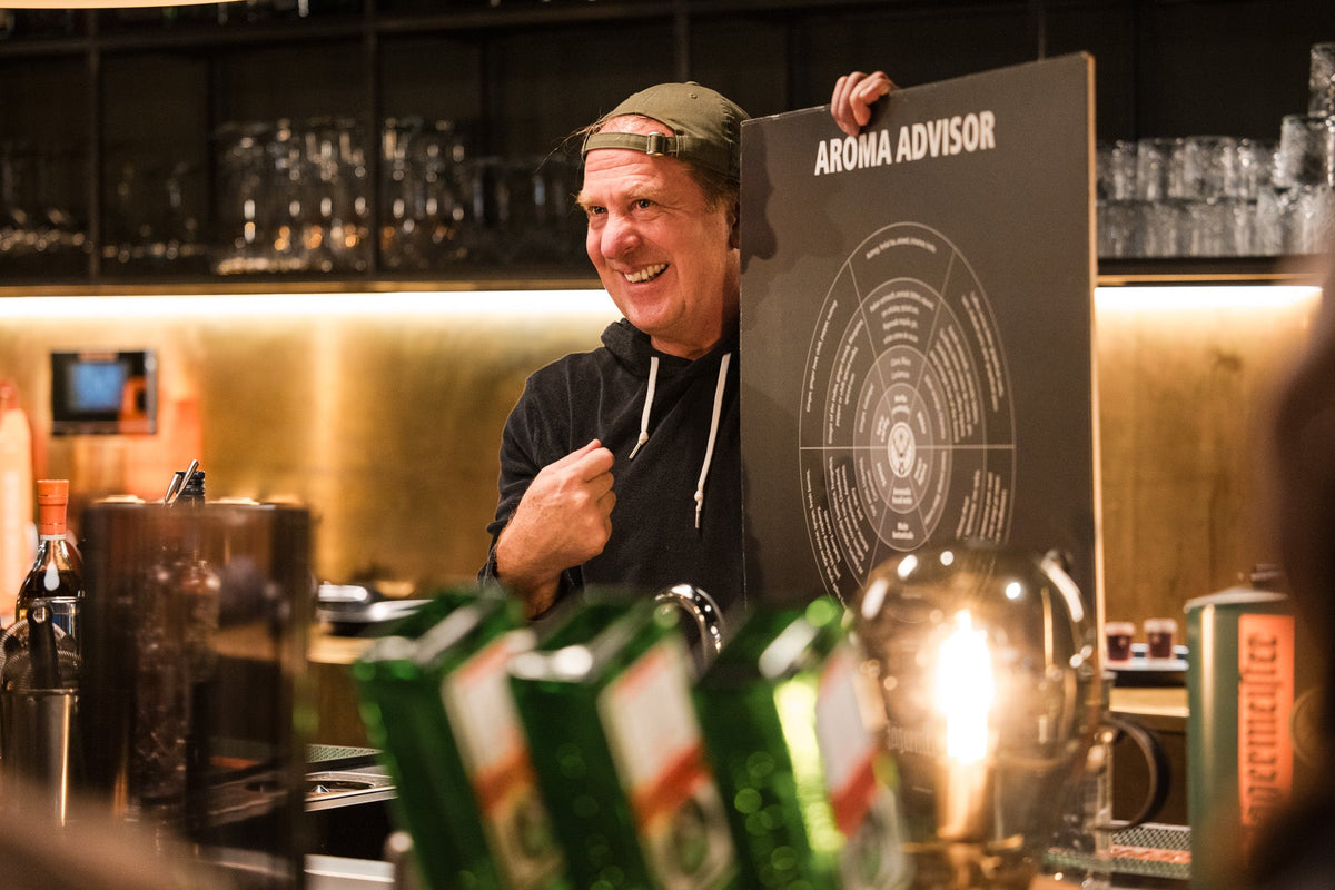 A man in a bar setting holds up a large black board labeled 'AROMA ADVISOR' with a circular scent wheel diagram. The scene includes bottles, glasses, warm lighting, and blurred foreground elements.