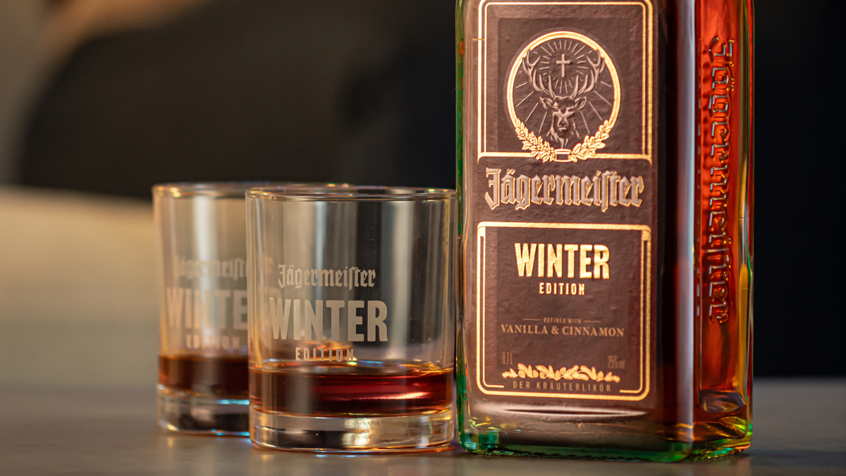 A bottle of Jägermeistetter Winter Edition liquor next to two clear glasses filled with the amber liquid, photographed on a tabletop with a softly blurred indoor background.
