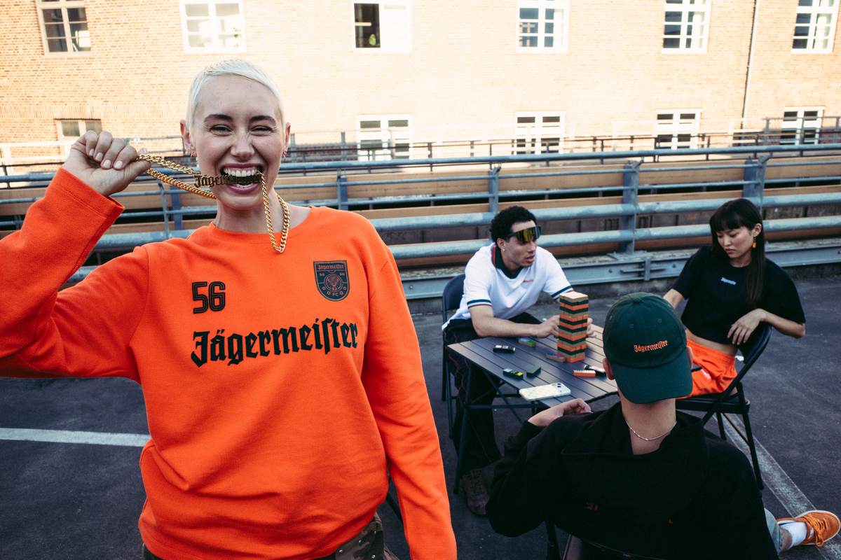 A smiling woman in the foreground wears a bright orange long-sleeve sweatshirt with '56 Jägermeister' printed in black on the front, holding a Jägermeister gold chain. Three people are seated at a table in the background on an outdoor rooftop or deck, with a brick building behind them.