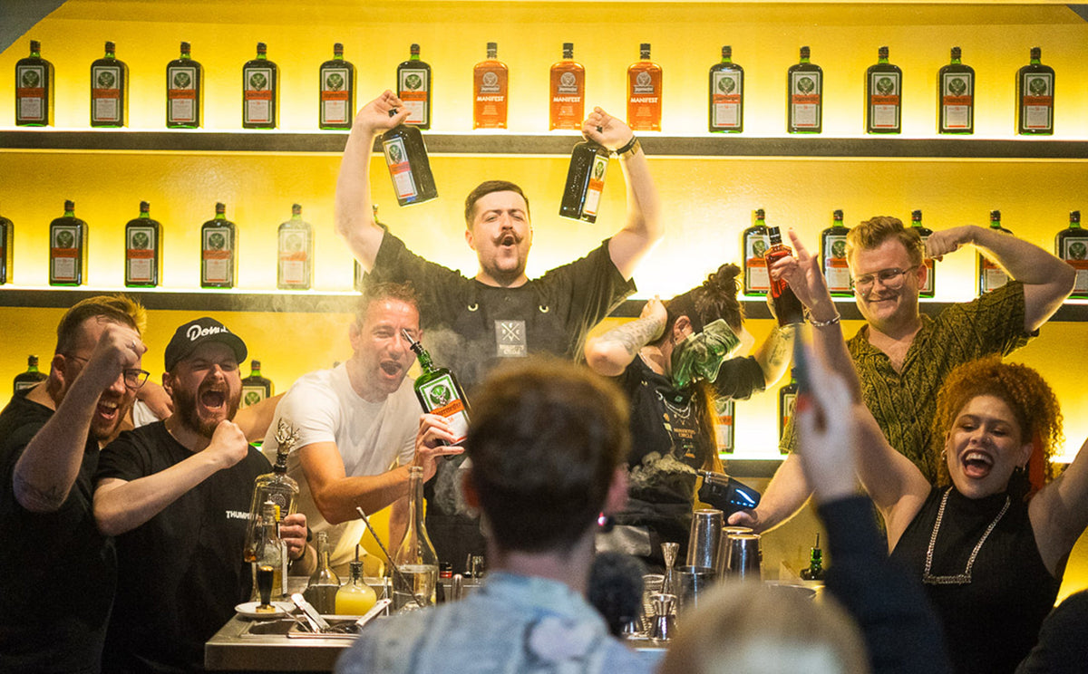 Group of people standing behind a bar counter, raising bottles and drinks toward the camera, with shelves of illuminated bottles lining a yellow backlit wall behind them.
