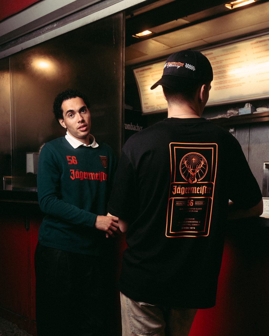Two men standing outside a red food truck at night. One man faces the camera wearing a dark green Jägermeister Longsleeve and black pants. The other one is wearing the Jägermeister Racing Cap and the Jägermeister label t-shirt in black.