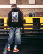 A model wearing streetwear apparel, including a black hoodie with the Jägermeister tradition graphic back print, standing on a dark paved surface in front of a brick building and a yellow train.