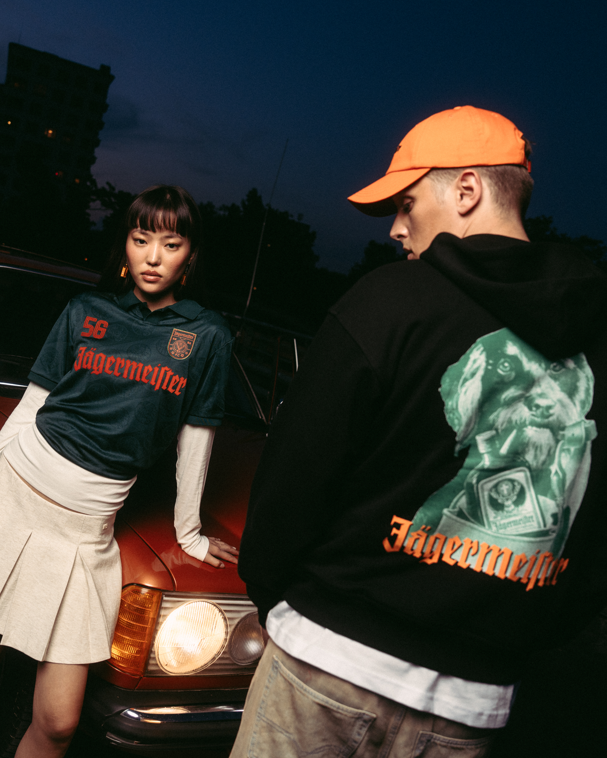 Two models posing in front of a vintage red car at dusk. One model wears a dark green Jägermeister jersey with a white pleated skirt, and the other wears a black Jägermeister Waldi hoodie and an orange Jägermeister Dad cap.