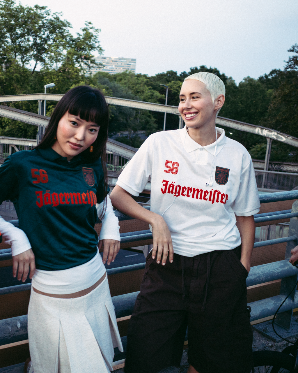 Quatro pessoas estão de pé ao ar livre numa ponte ou passadiço, vestidas com streetwear. Uma pessoa veste um casaco laranja vivo com fecho, outra veste uma camisola Jägermeister verde-escura com uma saia branca, uma terceira veste uma camisola Jägermeister branca com calções escuros, e uma quarta veste um hoodie preto e um boné laranja.