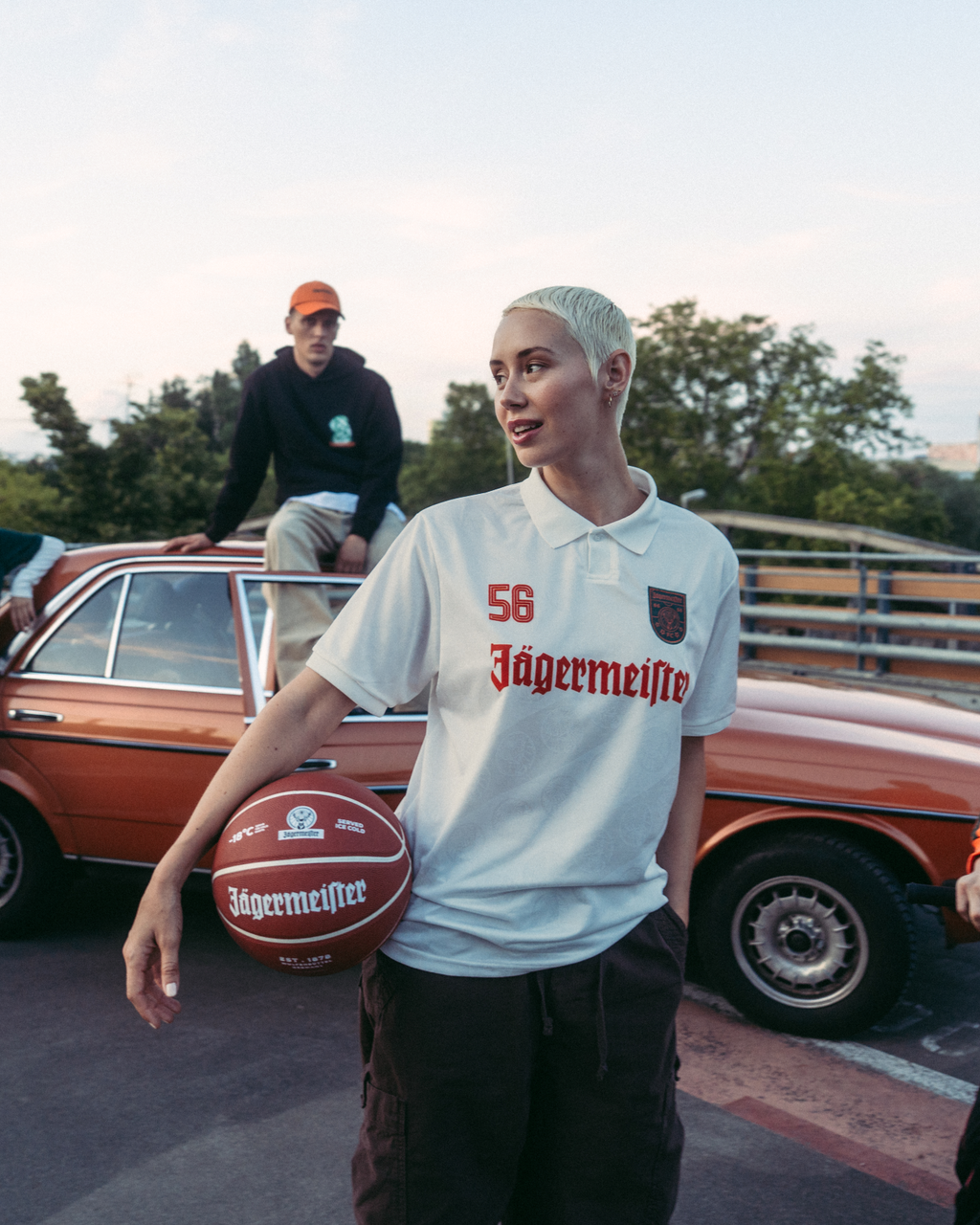 Um grupo de quatro pessoas a posar ao ar livre numa ponte ou passagem superior, com um carro laranja vintage atrás delas. A figura central segura uma bola de basquetebol e veste uma camisola Jägermeister branca, enquanto as restantes usam streetwear laranja vivo, verde e preto.