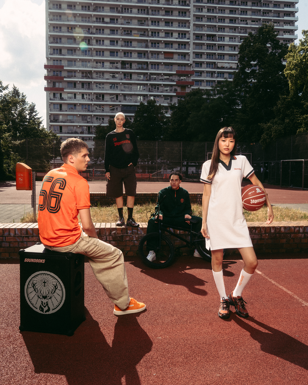 Cena de streetwear ao ar livre num campo de basquetebol com um edifício alto de apartamentos ao fundo. Um homem sentado com uma camisola Jägermeister laranja está sentado numa caixa preta parecida com uma coluna, enquanto uma mulher de pé com um vestido pólo branco Jägermeister segura uma bola de basquetebol.