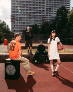 Cena de streetwear ao ar livre num campo de basquetebol com um edifício alto de apartamentos ao fundo. Um homem sentado com uma camisola Jägermeister laranja está sentado numa caixa preta parecida com uma coluna, enquanto uma mulher de pé com um vestido pólo branco Jägermeister segura uma bola de basquetebol.