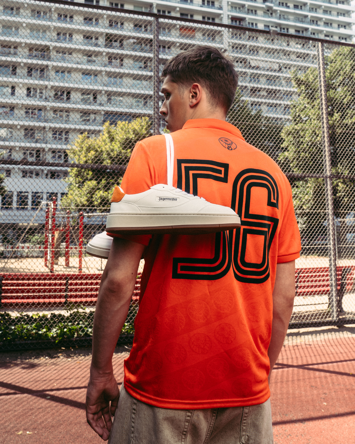 Uma pessoa está num campo desportivo ao ar livre com uma camisola Jägermeister laranja viva com o grande “56” impresso nas costas. Um par de ténis Jägermeister brancos está pendurado num ombro, com uma rede de arame e edifícios de apartamentos ao fundo.