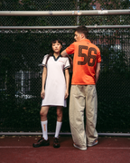 Dois modelos estão de pé num campo de ténis vermelho em frente a uma rede de arame. O modelo à esquerda veste um vestido pólo desportivo branco Jägermeister com detalhes em verde-escuro, meias brancas e sapatos pretos.