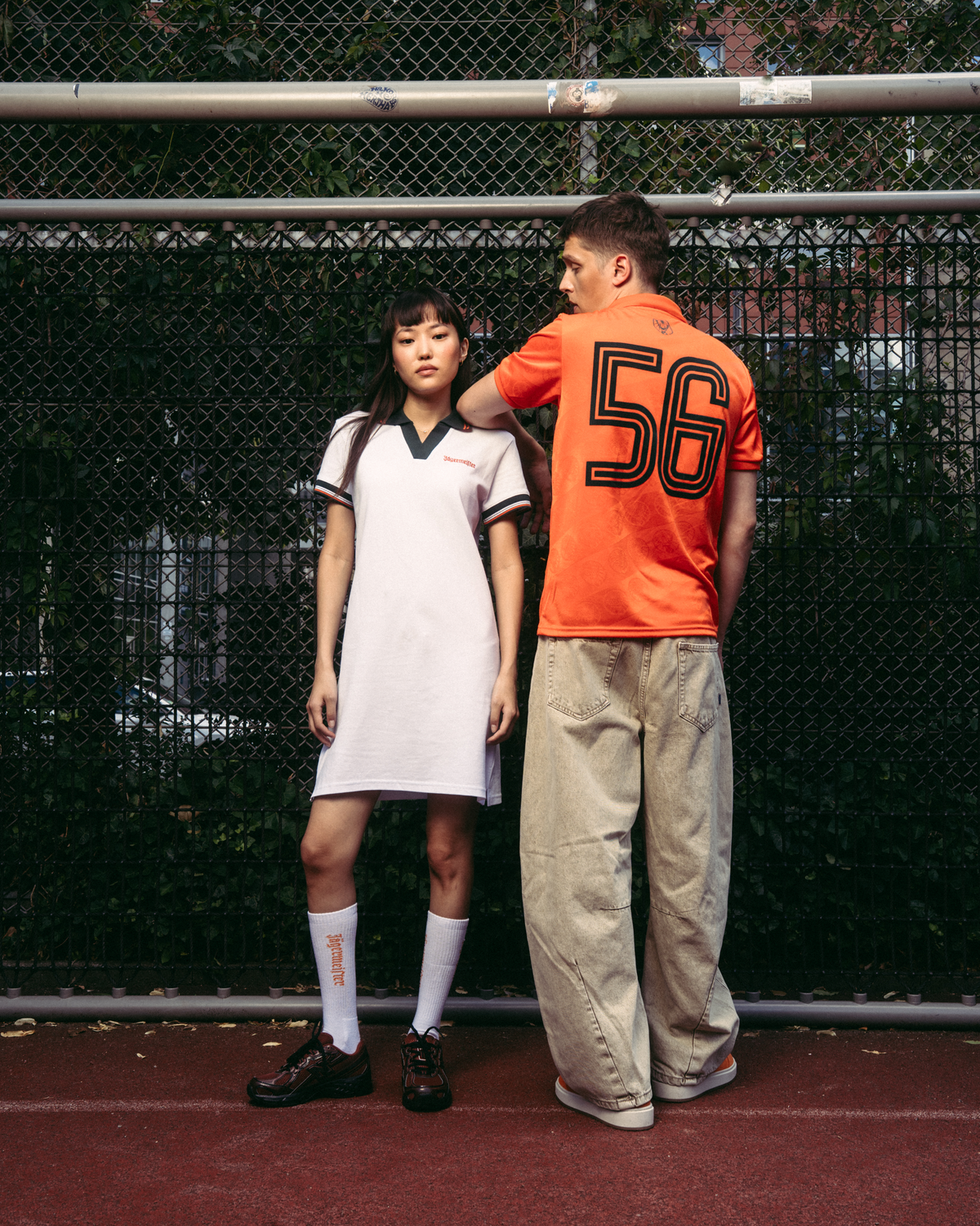 Dois modelos estão de pé num campo de ténis vermelho em frente a uma rede de arame. O modelo à esquerda veste um vestido pólo desportivo branco Jägermeister com detalhes em verde-escuro, meias brancas e sapatos pretos.