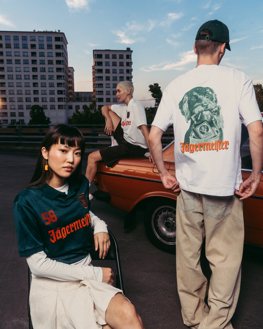 Três modelos com roupa com marca Jägermeister num cenário urbano ao ar livre, com um carro laranja vintage e edifícios de apartamentos ao fundo. Um modelo está sentado em primeiro plano com uma camisola Jägermeister verde-escura, outro está de costas para a câmara com uma T-shirt branca gráfica e calças bege, e um terceiro modelo está sentado no carro com uma T-shirt branca.
