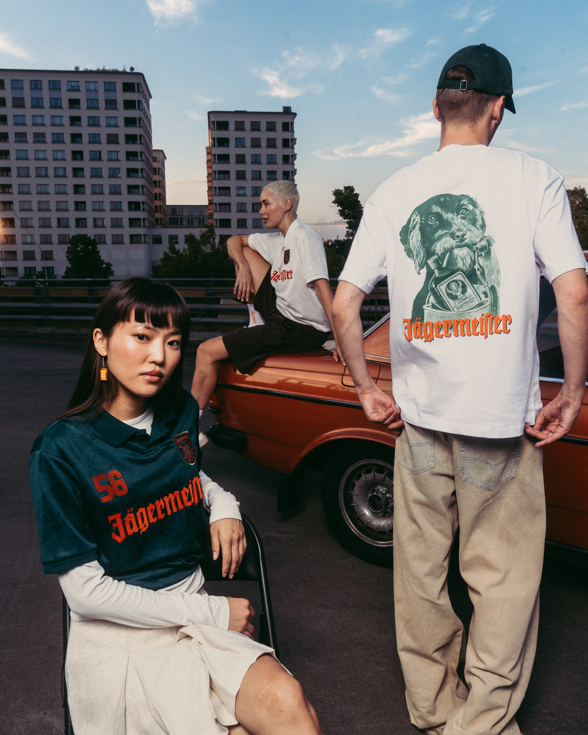 Três modelos com roupa com marca Jägermeister num cenário urbano ao ar livre, com um carro laranja vintage e edifícios de apartamentos ao fundo. Um modelo está sentado em primeiro plano com uma camisola Jägermeister verde-escura, outro está de costas para a câmara com uma T-shirt branca gráfica e calças bege, e um terceiro modelo está sentado no carro com uma T-shirt branca.