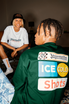Duas pessoas sentadas dentro de casa, uma com uma camisa branca com branding Jägermeister, a outra com um casaco verde com Jägermeister.