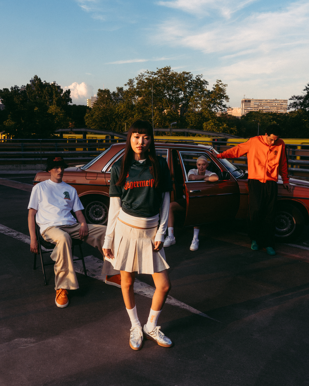 Um grupo de quatro jovens a posar ao ar livre numa pista de corrida ao pôr do sol, ao lado de um carro vermelho vintage. A figura central usa uma camisola Jäermeister verde-escura com letras laranja, uma saia branca plissada e ténis brancos.
