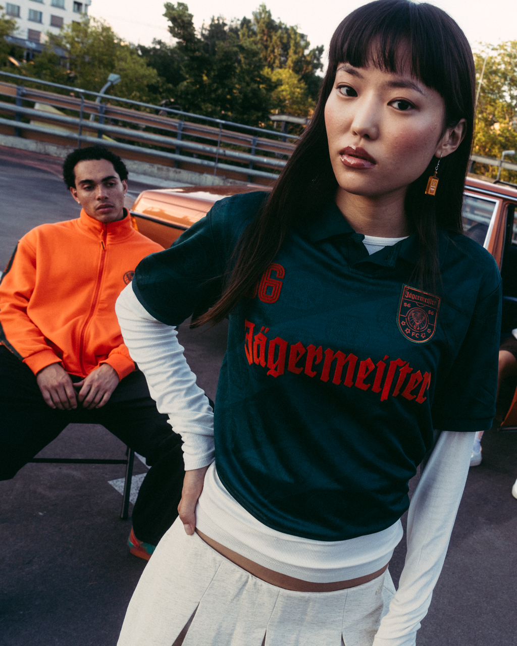Um grupo de quatro pessoas a posar ao ar livre em frente a um carro castanho vintage. A figura central usa uma camisola Jägermeister verde-escura, enquanto as restantes estão vestidas com streetwear casual.