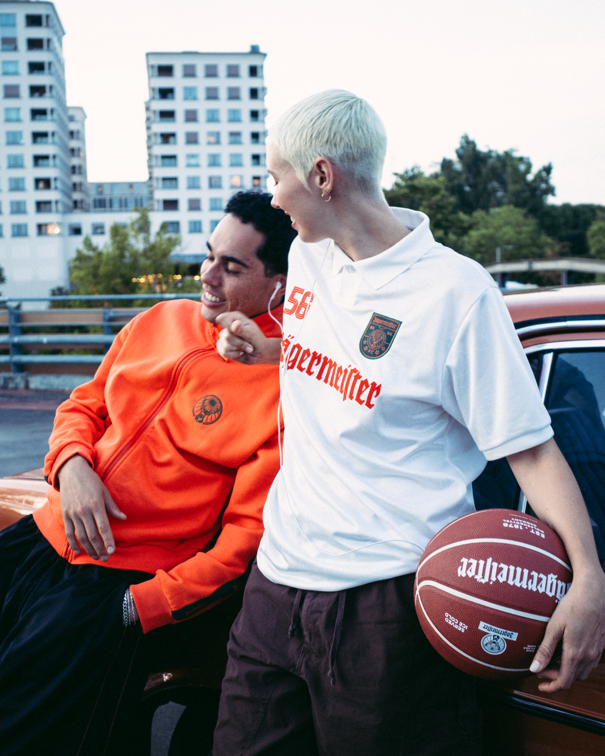 Two people standing beside a vintage red car in an outdoor parking area at dusk. One man wears a bright orange  Jägermeister sweatshirt and black pants, while the woman wears a white Jägermeister jersey and dark pants and holds a basketball.