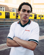 A man wearing a white Jägermeister short-sleeve polo shirt with dark green and orange trim, featuring the brand name 'Jägermeister' on the chest, standing with arms crossed in front of yellow trains and a brick building.