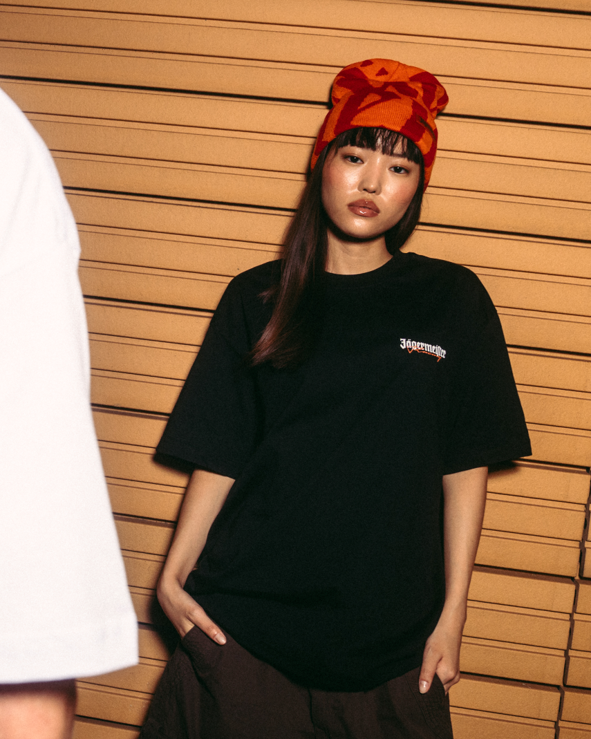 Two models posing against a wooden slat wall. The model on the left shows the back of a white t-shirt with a large orange graphic print reading 'Jägermeister'.