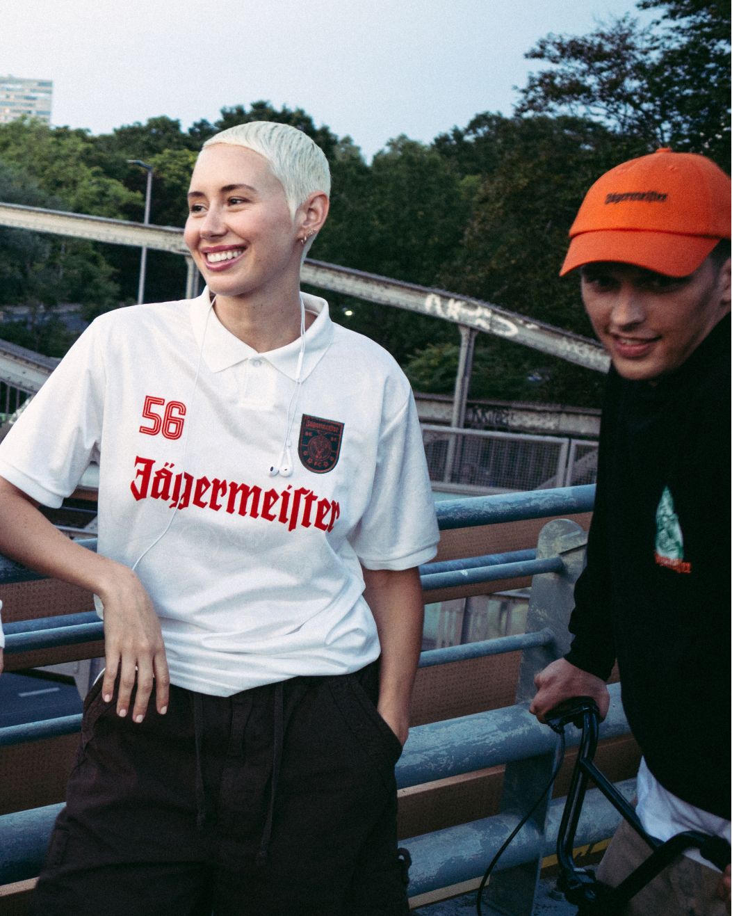 Four people standing outdoors on a bridge or walkway, wearing Jägermeister streetwear. One person wears a bright orange zip-up jacket, another wears a dark green Jagermeister jersey with a white skirt, a third wears a white Jagermeister jersey with dark shorts, and a fourth wears a black Jägermeister hoodie and an orange cap.