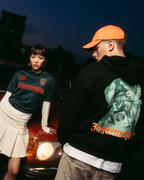 Duas modelos a posar em frente a um carro vermelho vintage ao anoitecer. Uma modelo veste uma camisola verde-escura 'Jägermeister' com uma saia branca pregueada, e a outra veste um hoodie preto Jägermeister Waldi e um boné laranja.