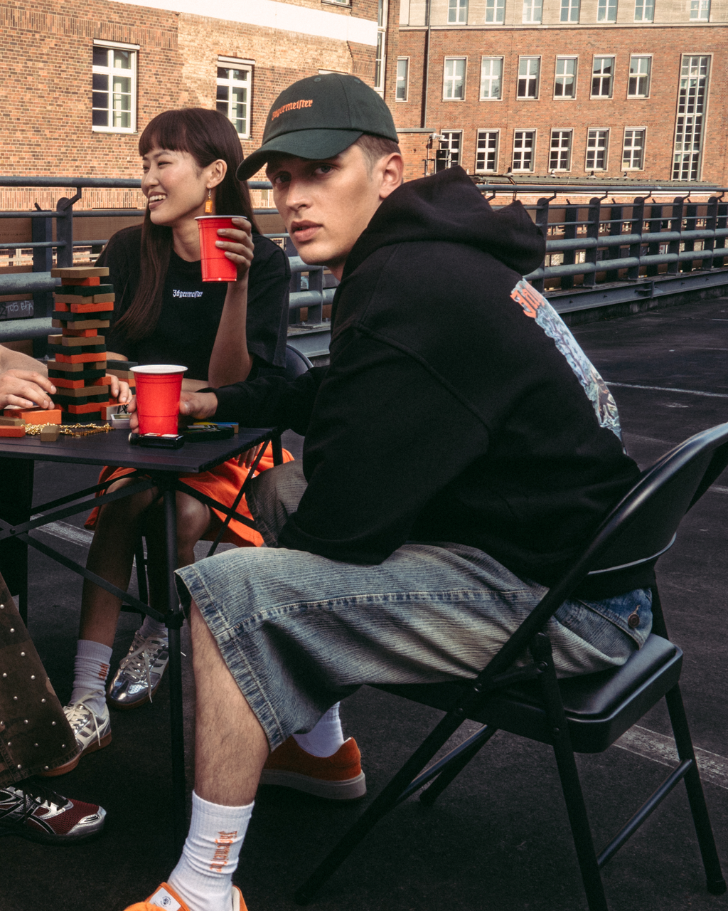 Quatro pessoas sentadas à volta de uma pequena mesa num terraço exterior ou numa ponte, a segurar copos vermelhos. Vestem streetwear casual Jägermeister, incluindo uma sweatshirt laranja, uma t-shirt branca, um hoodie preto e calças com padrão.