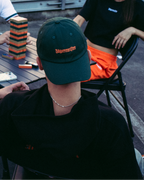 Uma mulher sorridente em primeiro plano usa uma sweatshirt de manga comprida laranja-viva com '56 Jägermeister' estampado a preto na frente, enquanto segura uma corrente dourada. Três pessoas estão sentadas a uma mesa ao fundo, num terraço ou deck exterior, com um edifício de tijolo atrás delas.