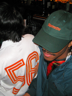 Duas pessoas sentadas juntas num ambiente de bar, uma delas com um boné tipo dad verde Jägermeister.