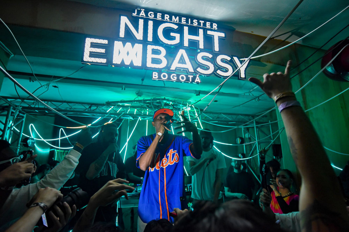 A person in a blue and orange baseball-style shirt stands in a crowded indoor venue under a large neon sign reading 'JÄGERMEISTER NIGHT EMBASSY BOGOTÁ', surrounded by people and glowing teal neon lines.