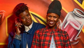 Two young men posing in front of a colorful graffiti-style wall. The man on the left wears a blue denim jacket over a white shirt, holding a phone to his ear.