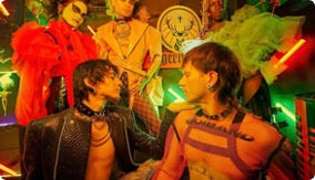 A group of people in elaborate costume and theatrical makeup in a brightly lit club or performance space with neon green and red lighting.