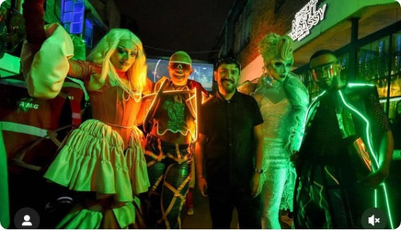 A group of five people posing together in a dimly lit, neon-lit venue at night. Several individuals are wearing elaborate costumes with glowing neon accents, including a blonde figure in a light green dress, a figure in a metallic silver outfit, and a figure in black with green neon lines.