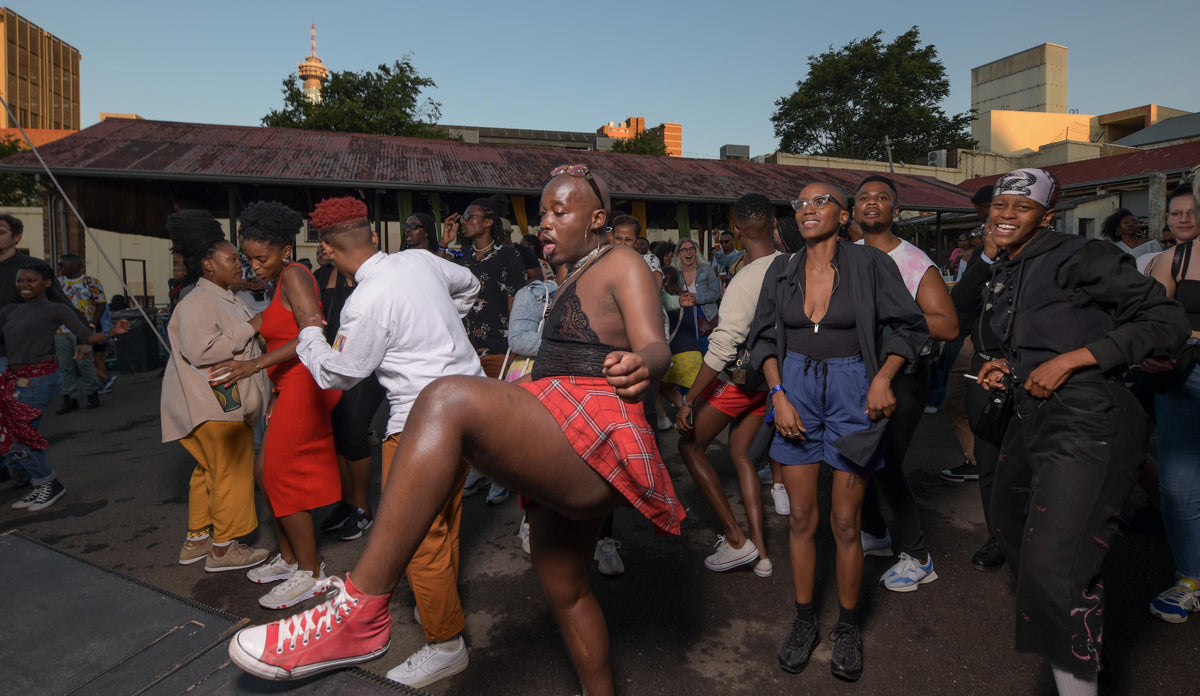 A crowd of people dancing outdoors in front of a building at dusk, with a central person in a black top and red plaid skirt raising one leg.