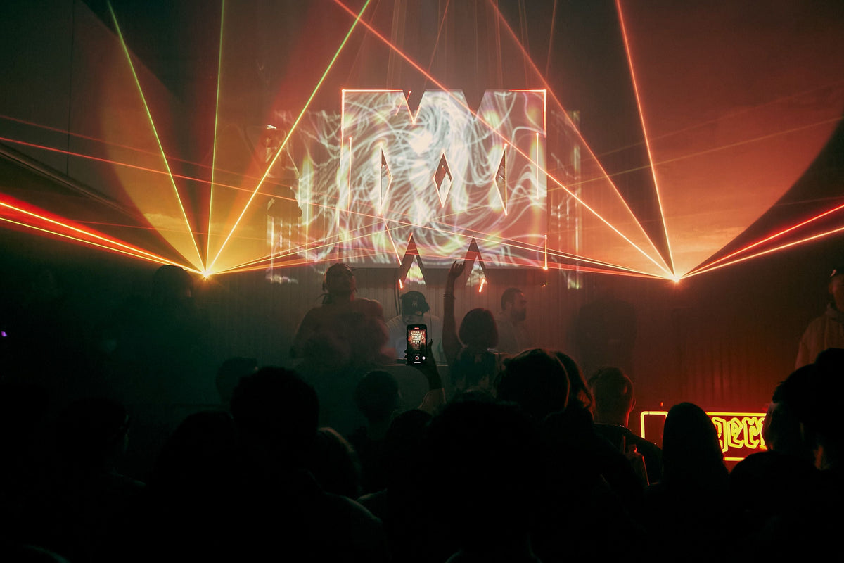 A dimly lit nightclub or event space with a crowd of people in the foreground, red and orange laser beams sweeping across the room, and a large illuminated screen displaying abstract light patterns of the Night Embassy logo in the background.