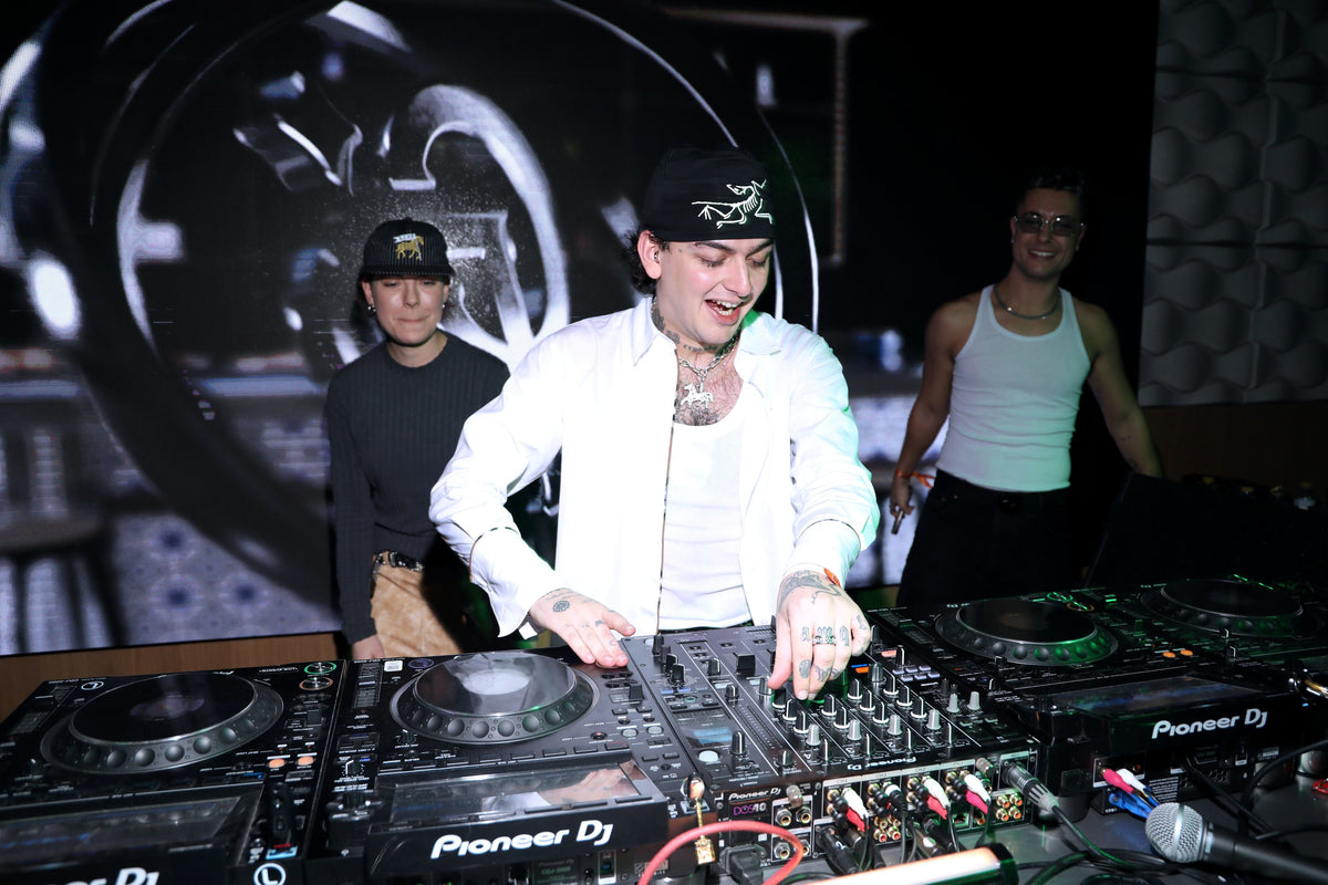 A DJ in a white shirt and black cap is operating a Pioneer DJ turntable setup in a dark venue, with two other men standing behind him.