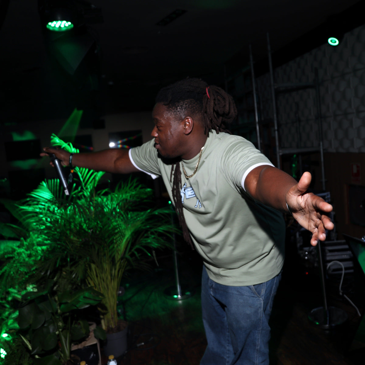 A man with dreadlocks wearing a light sage green t-shirt and blue jeans stands with arms outstretched in a dimly lit venue with green stage lighting.