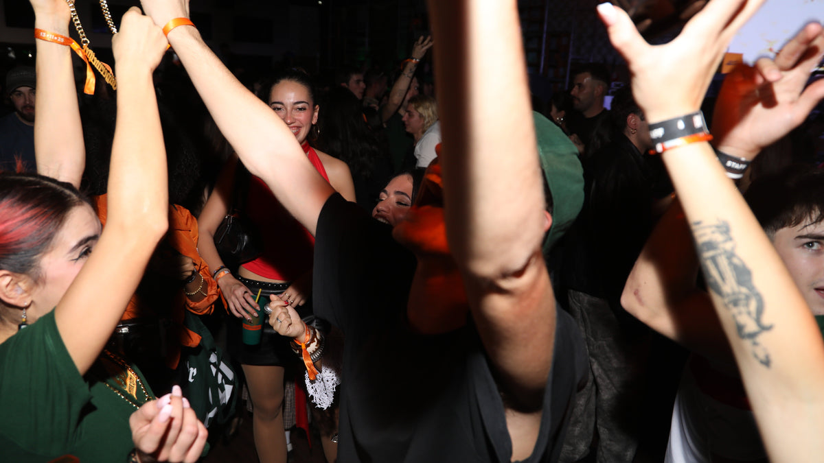 A crowded nightclub or concert scene with multiple people raising their arms, wearing wristbands, and illuminated by low light with strong orange and red lighting.