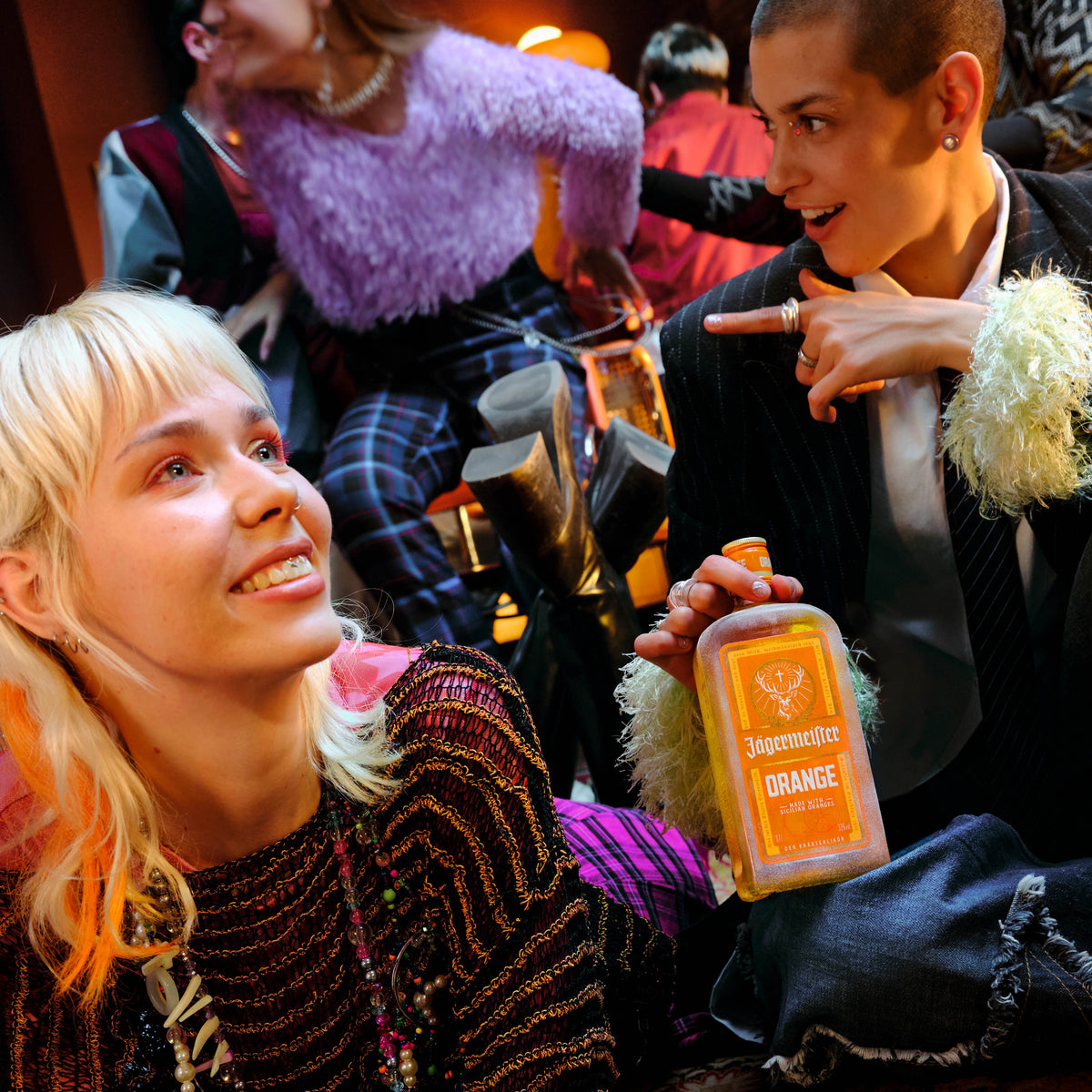 A person in a black pinstripe jacket with a white feather collar holds a bottle of Jägermeister Orange Liqueur while smiling at another person in a party setting.
