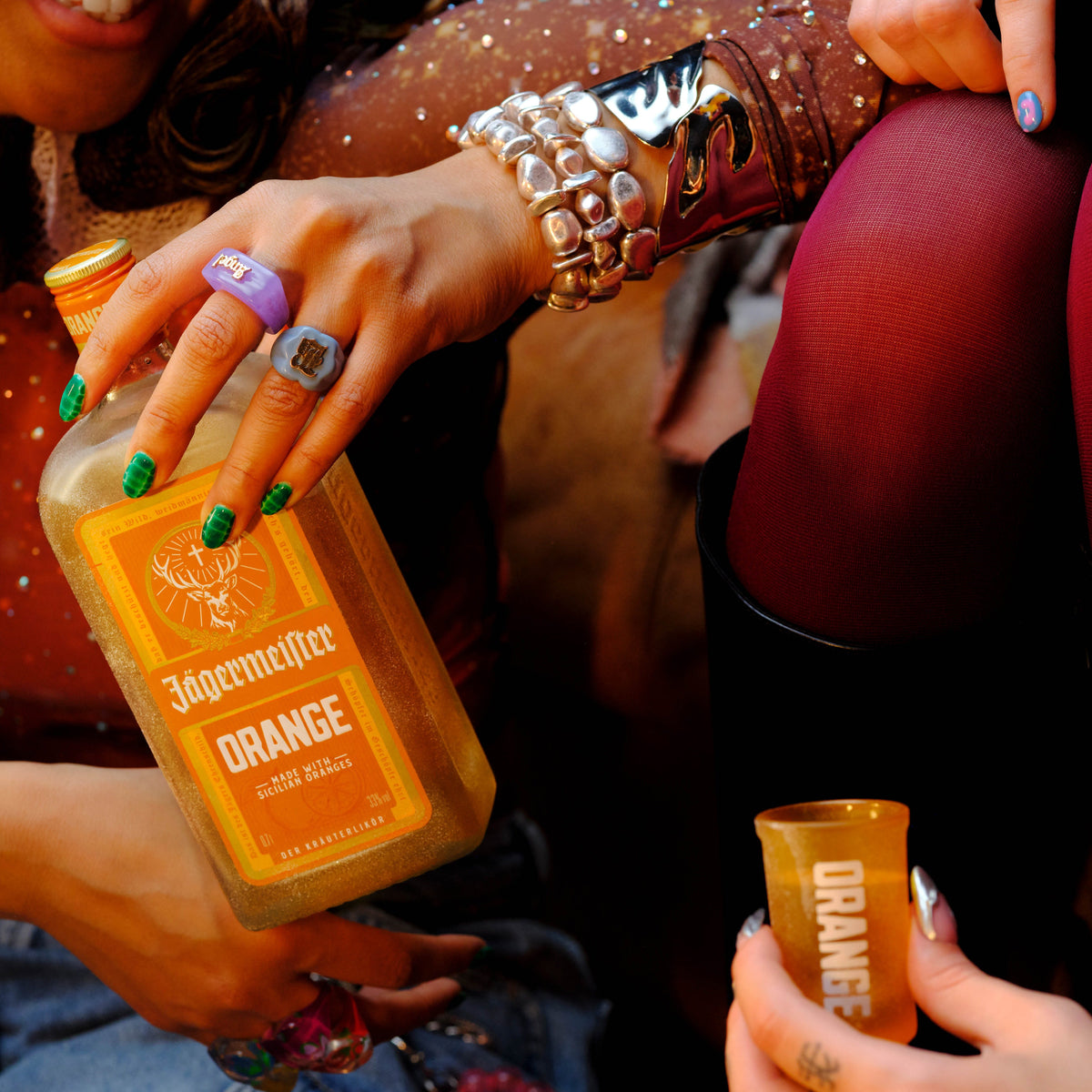 A close-up lifestyle shot of a person holding a bottle of Jägermeister Orange flavored tincture. The person is wearing a gold bracelet, a purple ring, and green nail polish, and is holding an orange shot glass.