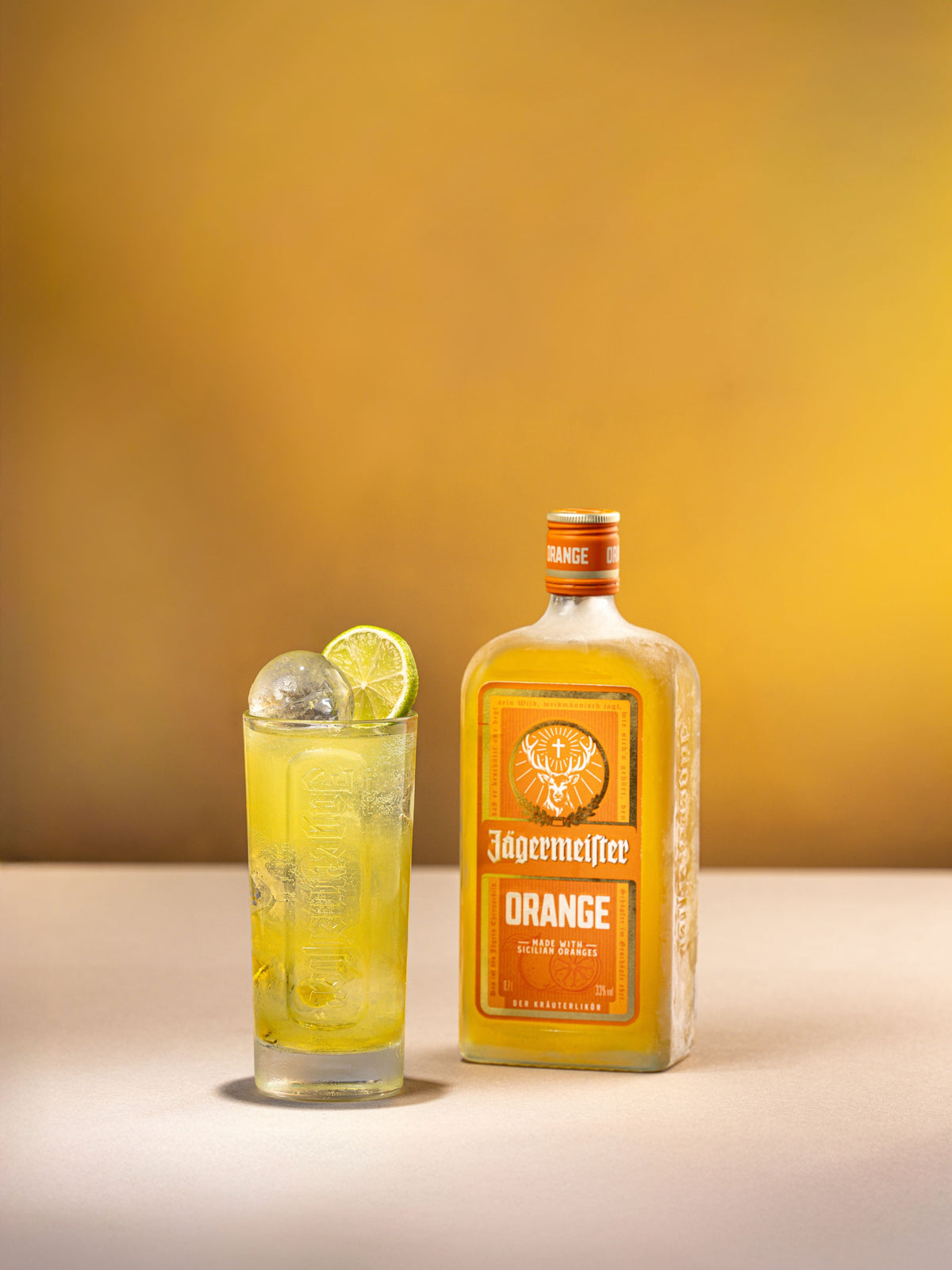 A bottle of Jagermeister Orange liqueur stands next to a tall glass filled with a yellow-orange cocktail and ice, garnished with a lime wheel, against a warm golden background.