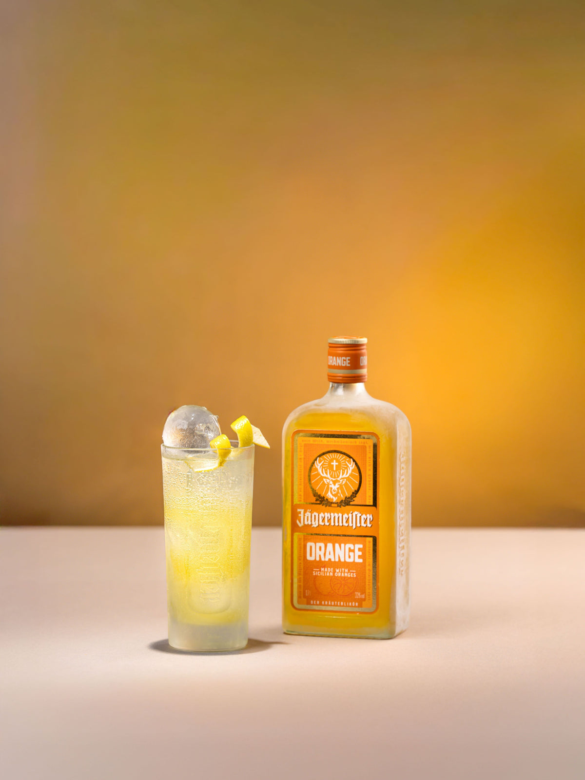 A bottle of Jägermeier Orange liqueur stands on a light surface beside a tall glass filled with a pale yellow drink, ice, and a small orange peel garnish, against a warm yellow-to-brown gradient background.