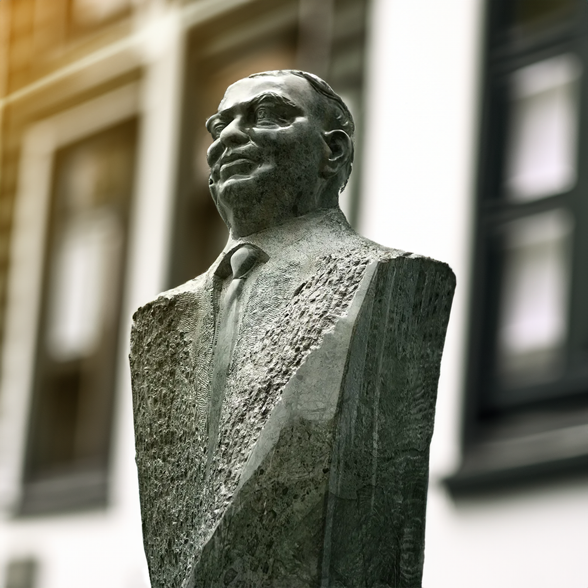 A stone bust sculpture of a man in a suit and tie, photographed outdoors against a softly blurred building facade.