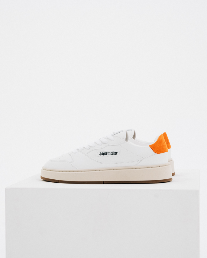 A white Jägermeister sneaker by with a bright orange heel tab and black logo text on the side, displayed on a white pedestal against a white background.