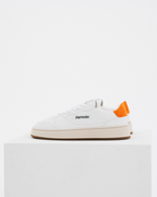 A white Jägermeister sneaker by with a bright orange heel tab and black logo text on the side, displayed on a white pedestal against a white background.