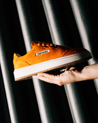 A hand holds an orange Jägermeister sneaker with a white sole and a visible 'Jägermeister' label, set against a dark background with vertical metallic slats.