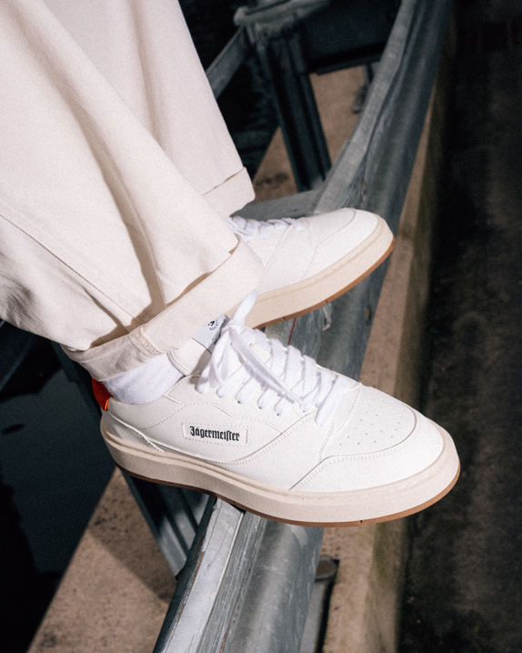 A person wearing off-white Jägermeister sneakers with white socks and light beige pants, sitting on a metal railing. The shoes feature a white leather upper, white laces, a gum sole, and a small black 'Jägermeister' logo on the side.