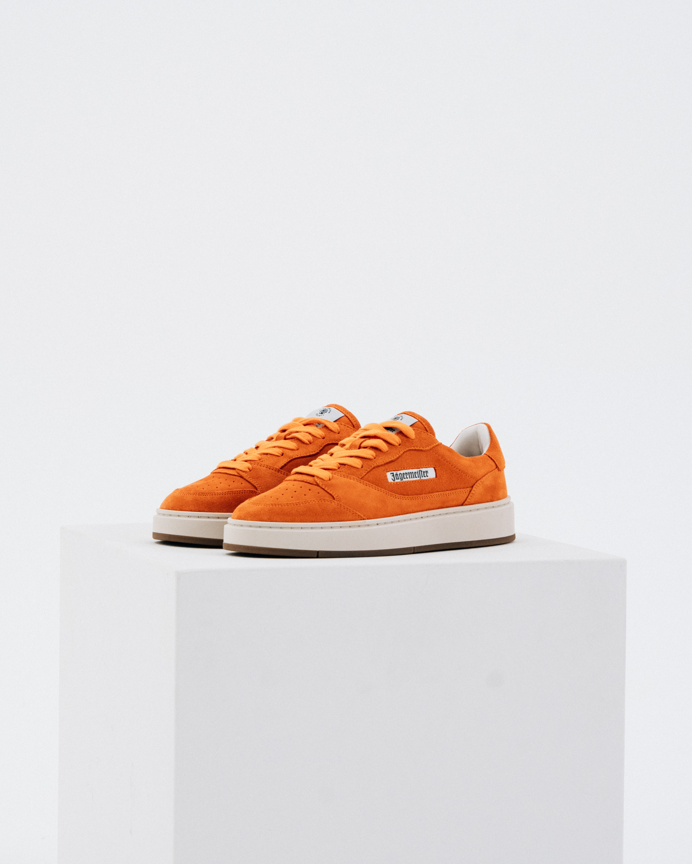 A pair of orange Jägermeister sneakers with white midsoles and gum soles, displayed on a white geometric pedestal against a light gray background.