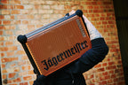 Person holding an orange and black portable Soundboks speaker with 'Jägermeister' branding in front of a brick wall.