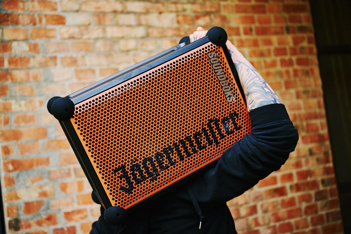 Person holding an orange and black portable Soundboks speaker with 'Jägermeister' branding in front of a brick wall.