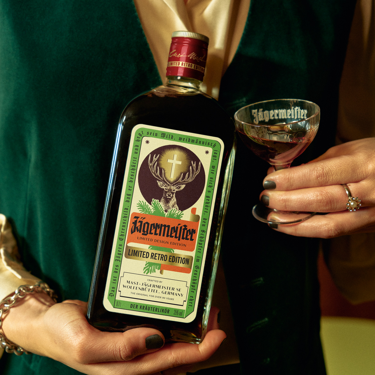 A person in a dark green velvet jacket holds a bottle of Jägermeister Limited Retro Edition a stemmed glass with the Jägermeister logo.