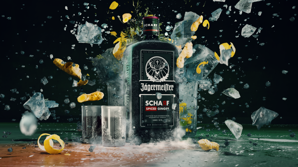 A bottle of Jägermeier Schaf stands in the center of a dramatic splash scene with ice cubes, liquid, and lemon peels flying around it against a dark background.